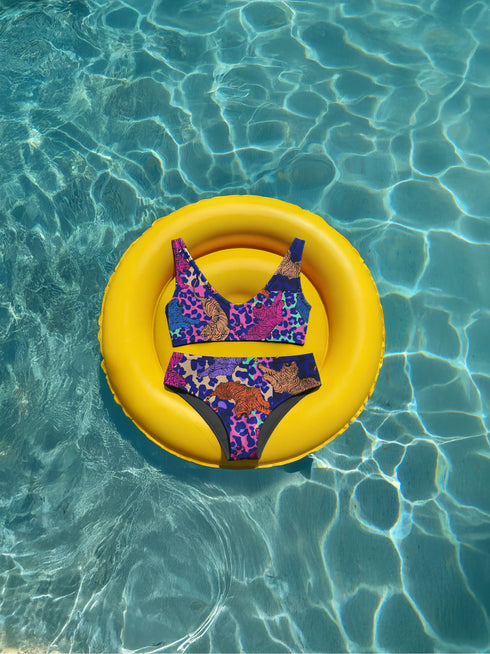 Psychedelic animal print bikini top with multicolor tiger pattern in neon pink, purple, orange, and blue, made of stretchy recycled fabric with wide shoulder straps and scoop neckline.